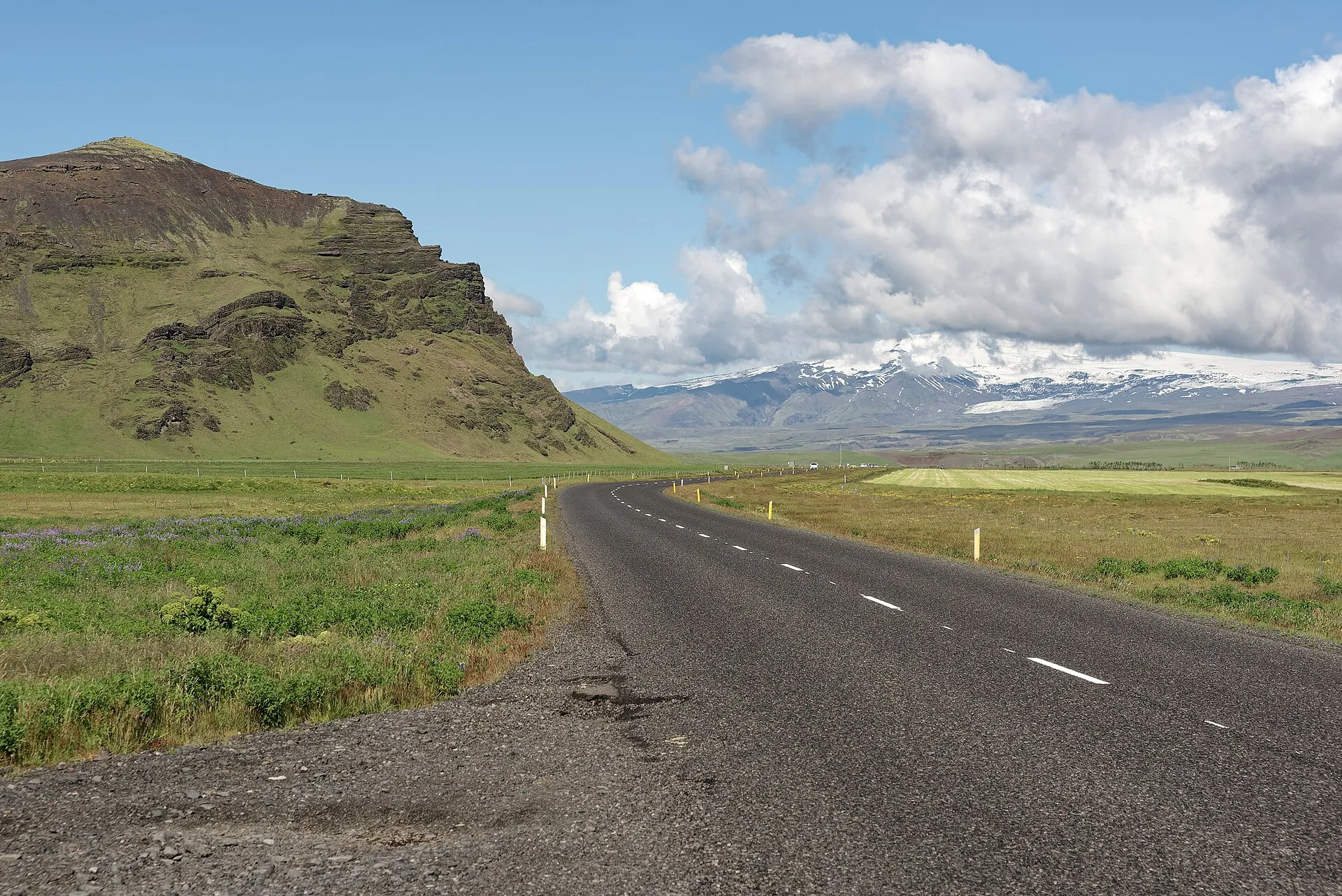 Asphaltierte Ringstraße in Südisland zwischen Wiesen, links ein Felsberg, hinten der verschneite Gletscher Mýrdalsjökull