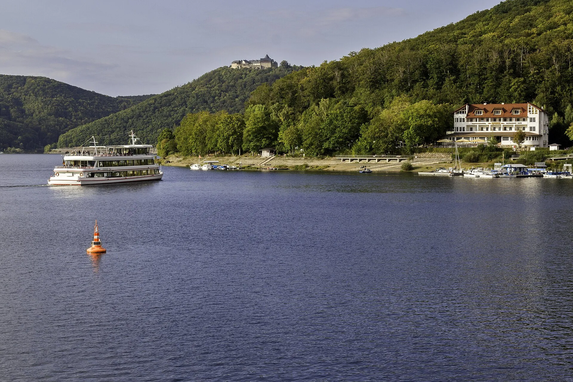 Ausflugsschiff auf dem Edersee, darüber auf dem bewaldeten Berg Schloss Waldeck, rechts ein Hotel am Ufer