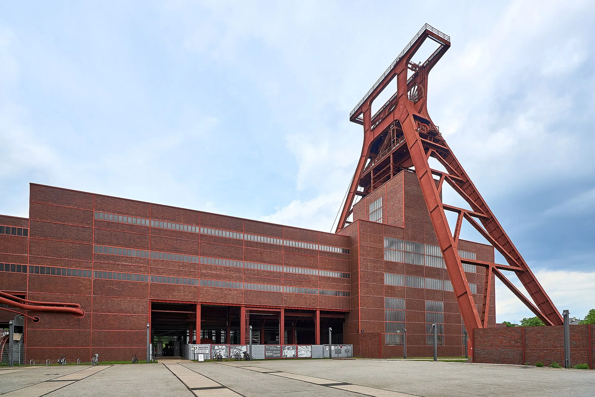 Rotes Fördergerüst über Schacht XII der Zeche Zollverein neben dem Backsteinbau, darüber bewölkter Himmel